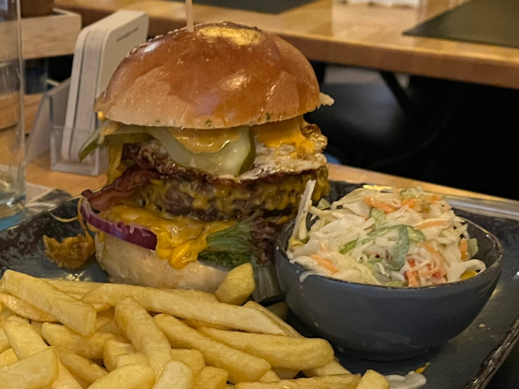 Das Bild zeigt einen Bildausschnitt: Burger, Pommes und Salat. Ober- und unterhalb des Motivs ist jeweils ein schmaler weißer und ein breiter blaugrüner Streifen. Oben rechts ist das Logo von BM 3X21, unten links ein QR-Code und unsere Kontaktdaten: bm3x21@web.de | www.bm3x21.de