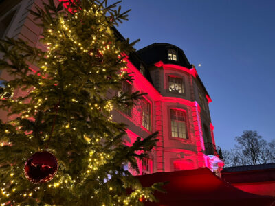 In der Bildmitte sieht man einen Anschnitt des rot angestrahlten Schloss Türnich. Ein Teil des Schlosses wird durch einen geschmückten Weihnachtsbaum am linken Bildrand verdeckt.