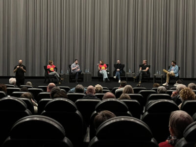 In der Bildmitte sitzen sieben Menschen nebeneinander auf einer Bühne vor einem grauen Theatervorhang. Im Vordergrund sieht man die Hinterköpfe zahlreicher Kinobesucherinnen und -besucher.