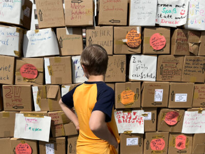 In der Bildmitte steht ein Junge mit einem orangefarbenen T-Shirt mit dem Rücken zur Kamera und betrachtet eine Mauer aus Pappkartons. Auf die Kartons sind Bildungshemmnisse geschrieben.