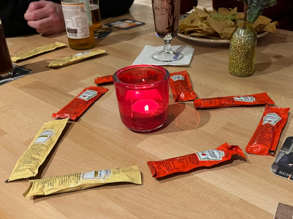 Ein Teelicht in einem roten Teelichtglas steht auf einer Holztischplatte. Um das Glas herum liegt ein aus mehreren Ketchup- und Majo-Portionsbeutel geformtes Herz. Im Hintergrund sind halb ausgetrunkenen Gläser mit verschiedenen Getränken und ein Teller mit Nachos zu sehen.