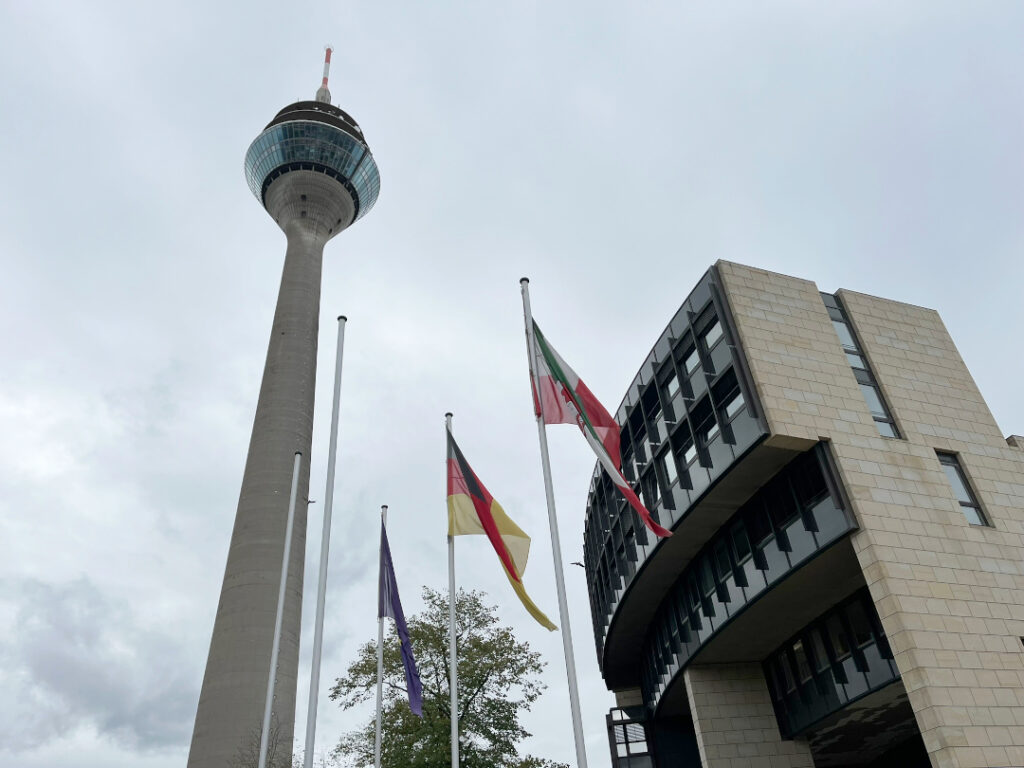 Vor einem wolkenverhangenen Himmel sind der Düsseldorfer Fernsehturm, ein Teil des Landtags NRW und drei Fahnen zu sehen: Europa, Deutschland, NRW.