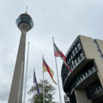 Vor einem wolkenverhangenen Himmel sind der Düsseldorfer Fernsehturm, ein Teil des Landtags NRW und drei Fahnen zu sehen: Europa, Deutschland, NRW.