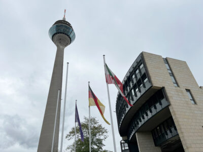 Vor einem wolkenverhangenen Himmel sind der Düsseldorfer Fernsehturm, ein Teil des Landtags NRW und drei Fahnen zu sehen: Europa, Deutschland, NRW.
