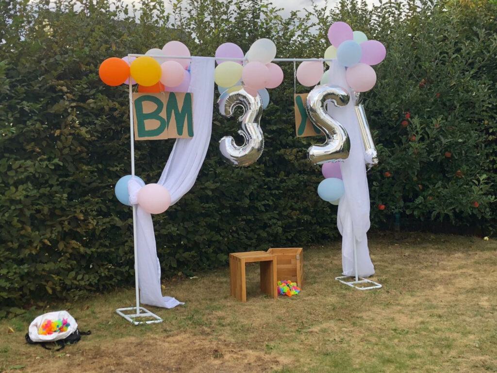 Unser Selfiespot: Vor einer Hecke steht ein Ständer aus zwei senkrechten und einer horizontalen Stange vor einer Hecke. An der horizontalen Stange hängen bunte Luftballons und BM 3X21 aus blaugrünen Buchstaben aus Pappe und großen silbernen Zahlenluftballons. Links und rechts hängen weiße Vorhangschals herunter.