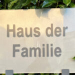 Auf einem silbernen Schild steht in grauer Schrift: Haus der Familie. Hinter dem Schild ist eine saftig grüne Hecke.