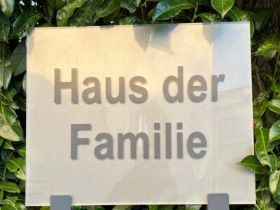 Auf einem silbernen Schild steht in grauer Schrift: Haus der Familie. Hinter dem Schild ist eine saftig grüne Hecke.