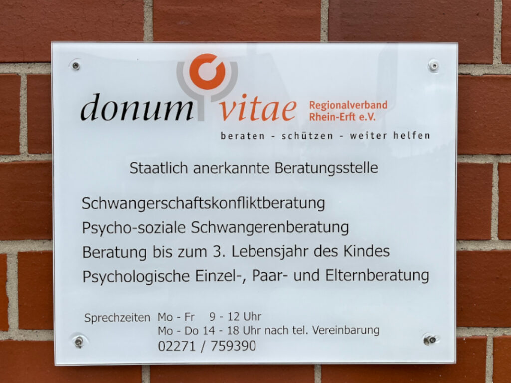 An einer roten Ziegelmauer hängt ein Schild. Auf diesem steht: „donum vitae Regionalverband Rhein-Erft e.V.. beraten – schützen – weiter helfen. Staatlich anerkannte Beratungsstelle. Schwangerschaftskonfliktberatung. Psycho-soziale Schwangerenberatung. Beratung bis zum 3. Lebensjahr des Kindes. Psychologische Einzel-, Paar- und Elternberatung. Sprechzeiten Mo bis Fr 9 bis 12 Uhr. Mo bis Do 14 bis 18 Uhr nach telefonischer Vereinbarung. 02271 759390.
