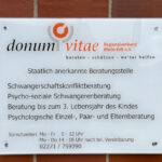 An einer roten Ziegelmauer hängt ein Schild. Auf diesem steht: „donum vitae Regionalverband Rhein-Erft e.V.. beraten – schützen – weiter helfen. Staatlich anerkannte Beratungsstelle. Schwangerschaftskonfliktberatung. Psycho-soziale Schwangerenberatung. Beratung bis zum 3. Lebensjahr des Kindes. Psychologische Einzel-, Paar- und Elternberatung. Sprechzeiten Mo bis Fr 9 bis 12 Uhr. Mo bis Do 14 bis 18 Uhr nach telefonischer Vereinbarung. 02271 759390.