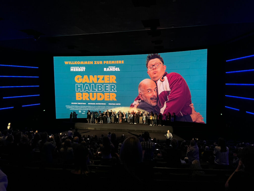 Blick durch einen dunklen Kinosaal mit vollen Rängen auf eine große Leinwand. Auf der Leinwand steht auf in weißer und gelber Schrift auf türkisblauem Hintergrund „Willkommen zur Premiere. Christoph Maria Herbst. Nicolas Randel. Ganzer halber Bruder.“ Rechts neben dem Text ist ein Foto der beiden Hauptcharaktere. Sie umarmen sich und haben dabei sehr lustige Gesichtsausdrücke zwischen Verzweiflung und Entsetzen. Vor der Leinwand stehen viele Menschen auf der Bühne, die an der Produktion beteiligt waren.