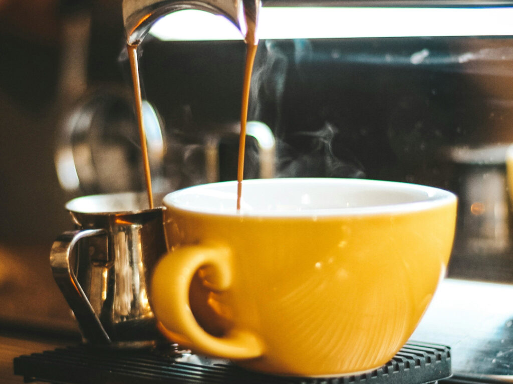 Eine gelbe Kaffeetasse und ein Edelstahl-Kännchen stehen unter dem 2-Wege-Aulauf einer Siebträger-Maschine.