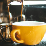 Eine gelbe Kaffeetasse und ein Edelstahl-Kännchen stehen unter dem 2-Wege-Aulauf einer Siebträger-Maschine.