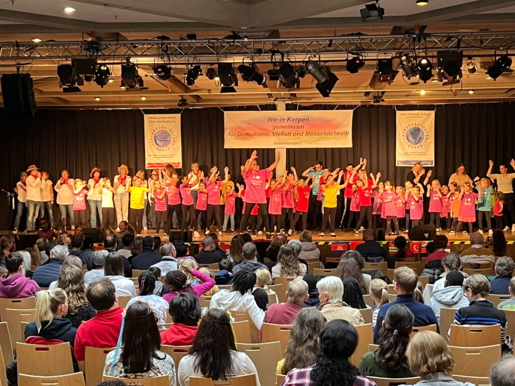 Im Vordergrund sieht man gut gefüllte Stuhlreihen von hinten. Die Besucher*innen schauen nach vorne auf die Bühne. Dort tanzen Kinder, Jugendliche und Erwachsene in schwarzen Hosen und bunten T-Shirts gemeinsam einen Tanz. Sie tanzen in mehreren Reihen hintereinander. In den vorderen Reihen wird mehr getanzt, in den hinteren mehr durch Klatschen supportet.