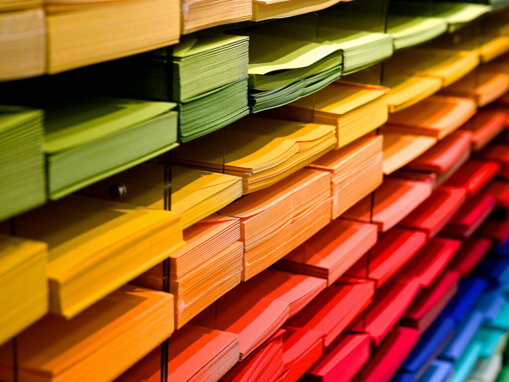 In der Bildmitte: Blick von schräg-vorne auf ein Regal mit Fächern mit Briefumschlägen und Briefpapier in alle Farben des Regenbogens.