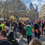 Die Teilnehmenden des Demozuges gehen den Neumarkt entlang. Es sind viele Menschen mit und ohne Behinderung zu sehen, die meisten von ihnen von hinten. Manche tragen große Blasinstrumente oder Transparente.