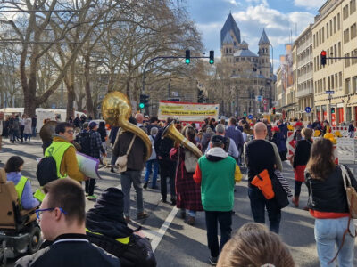 Die Teilnehmenden des Demozuges gehen den Neumarkt entlang. Es sind viele Menschen mit und ohne Behinderung zu sehen, die meisten von ihnen von hinten. Manche tragen große Blasinstrumente oder Transparente.