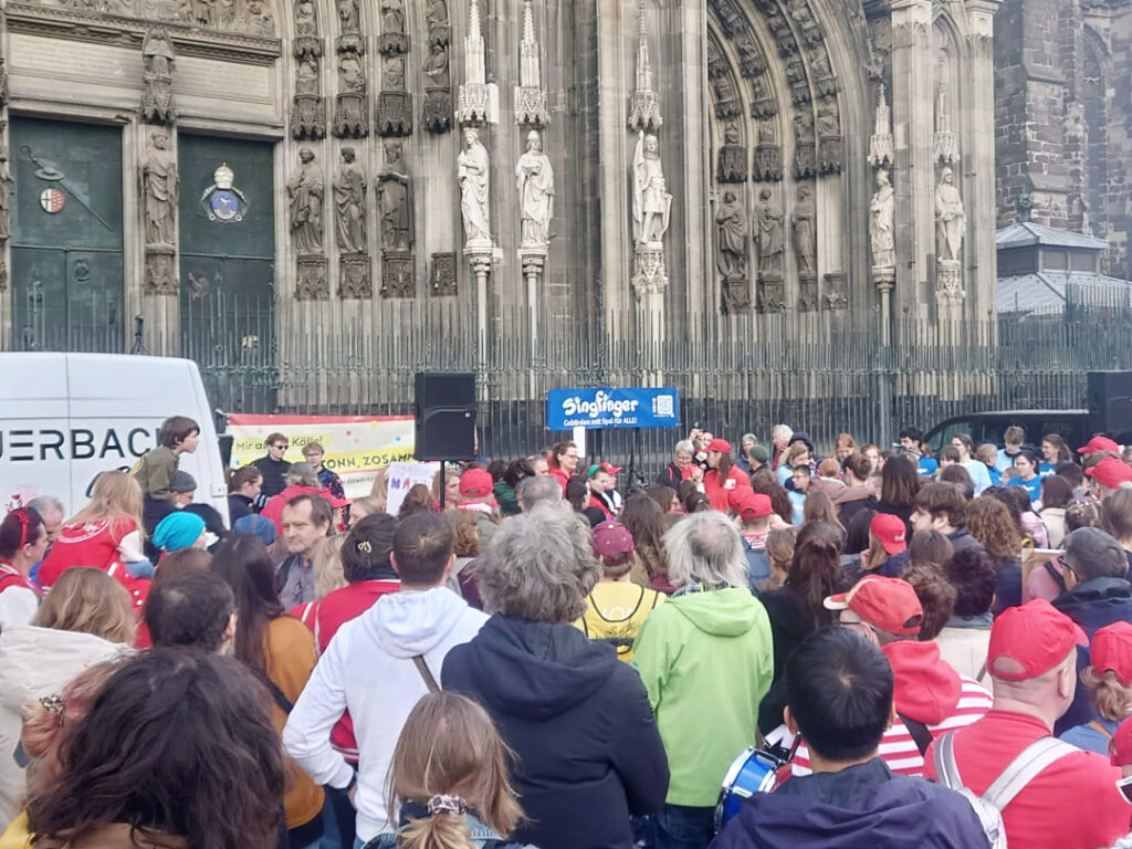 Kundgebung auf dem Roncalliplatz vor dem Kölner Dom. Es sind viele Menschen mit und ohne Behinderung zu sehen, die meisten von ihnen von hinten. Im Hintergrund sind Transparente von singfinger und mittendrin zu sehen, große Lautsprecher und der Bandbus von Auerbach.