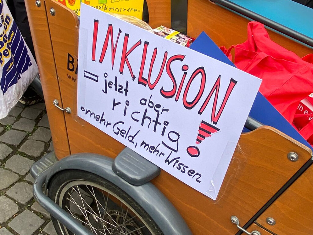 An einem Lastenrad hängt ein selbstgemaltes Plakat mit der Aufschrift: Inklusion - jetzt aber richtig! Mehr Geld, mehr Wissen