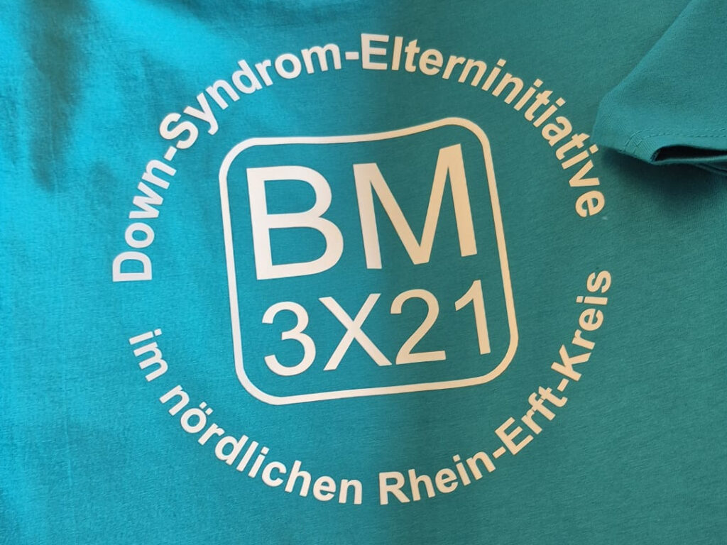 T-Shirt-Aufdruck BM 3X21: BM, darunter 3X21, beides umrandet von einem quadrat mit abgerundeten Ecken. In einem Kreis um dieses Logo herum der Schriftzug Down-Syndrom-Elterninitiative im nördlichen Rhhein-Erft-Kreis