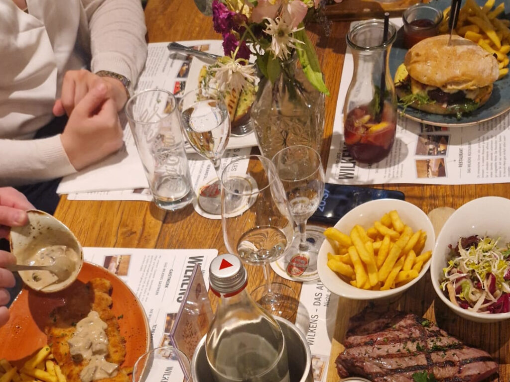 Blick von schräg oben auf einen Holztisch mit allerlei Getränken und Gerichten. Zu sehen sind unter anderem ein Jägerschnitzel, ein Brot mit Spargel, ein Burger und ein Steak mit Pommes.