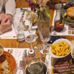 Blick von schräg oben auf einen Holztisch mit allerlei Getränken und Gerichten. Zu sehen sind unter anderem ein Jägerschnitzel, ein Brot mit Spargel, ein Burger und ein Steak mit Pommes.