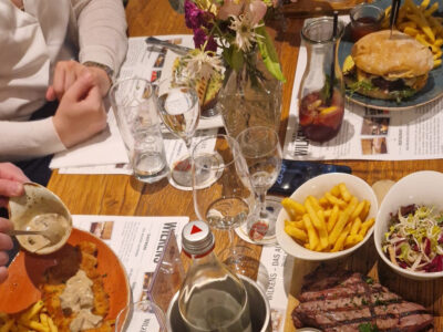 Blick von schräg oben auf einen Holztisch mit allerlei Getränken und Gerichten. Zu sehen sind unter anderem ein Jägerschnitzel, ein Brot mit Spargel, ein Burger und ein Steak mit Pommes.