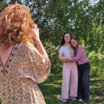 Fotografin von hinten. Sie hat rote Locken und trägt ein gemustertes Sommerkleid. Im Hintergrund stehen zwei Freundinnen mit Down-Syndrom im jungen Erwachsenenalter vor saftig grünen Bäumen.