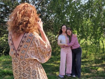Fotografin von hinten. Sie hat rote Locken und trägt ein gemustertes Sommerkleid. Im Hintergrund stehen zwei Freundinnen mit Down-Syndrom im jungen Erwachsenenalter vor saftig grünen Bäumen.