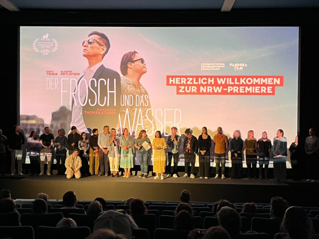 Blick durch den dunklen Kinosaal auf die helle Leinwand mit einem Standbild. Darauf die beiden Hauptdarsteller, ihre Namen der Titel des Films „Der Frosch und das Wasser“ und rechts daneben der Schriftzug Herzlich Willkommen zur NRW-Premiere. Vor der Leinwand stehen viele Menschen auf der Bühne, die bei der Produktion des Films mitgewirkt haben.