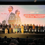 Blick durch den dunklen Kinosaal auf die helle Leinwand mit einem Standbild. Darauf die beiden Hauptdarsteller, ihre Namen der Titel des Films „Der Frosch und das Wasser“ und rechts daneben der Schriftzug Herzlich Willkommen zur NRW-Premiere. Vor der Leinwand stehen viele Menschen auf der Bühne, die bei der Produktion des Films mitgewirkt haben.
