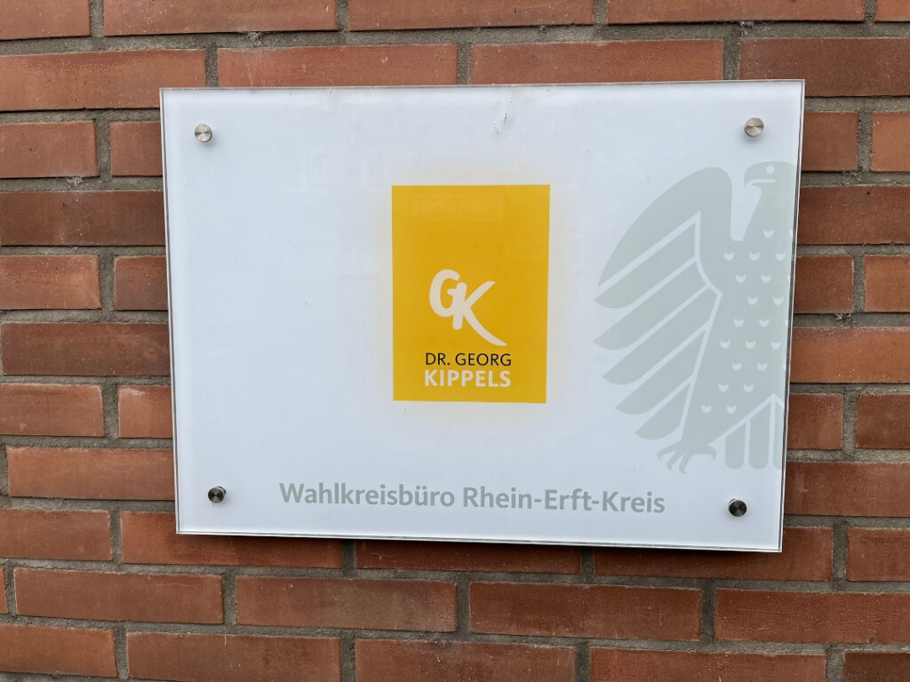 Glasschild im Querformat mit weißem Hintergrund. Darauf in der Mitte eine gelbe Box mit den Initialen GK und dem Schriftzug Dr. Georg Kippes. Unter der gelben Box steht in grauer Schrift „Wahlkreisbüro Rhein-Erft-Kreis“. Rechts neben der gelben Box ein halber Bundesadler in grau. Das Schild hängt an einer roten Backsteinwand.