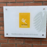 Glasschild im Querformat mit weißem Hintergrund. Darauf in der Mitte eine gelbe Box mit den Initialen GK und dem Schriftzug Dr. Georg Kippes. Unter der gelben Box steht in grauer Schrift „Wahlkreisbüro Rhein-Erft-Kreis“. Rechts neben der gelben Box ein halber Bundesadler in grau. Das Schild hängt an einer roten Backsteinwand.