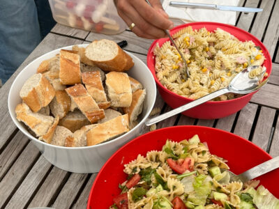 Ein Tisch mit zwei Nudelsalaten und einer großen Schüssel voller Baguettescheiben. Eine Männerhand tut sich Nudelsalat auf.