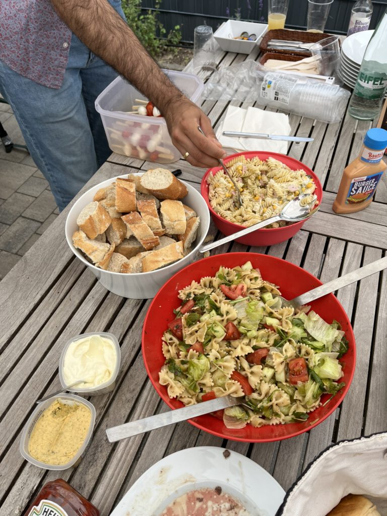 Ein Tisch mit zwei Nudelsalaten, einer großen Schüssel voller Baguettescheiben und anderen Leckereien. Eine Männerhand tut sich Nudelsalat auf.